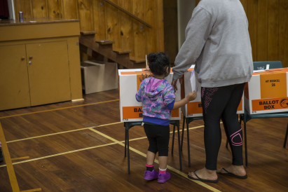 A child standing with an adult, as the adult puts their voting paper in a ballot box.